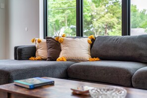 A grey sectional sofa with three cushions, a wooden coffee table with a book and a glass bowl, and a window with a view of trees. - Luxe Austin Riverside House w/ Skyline Deck (Austin)