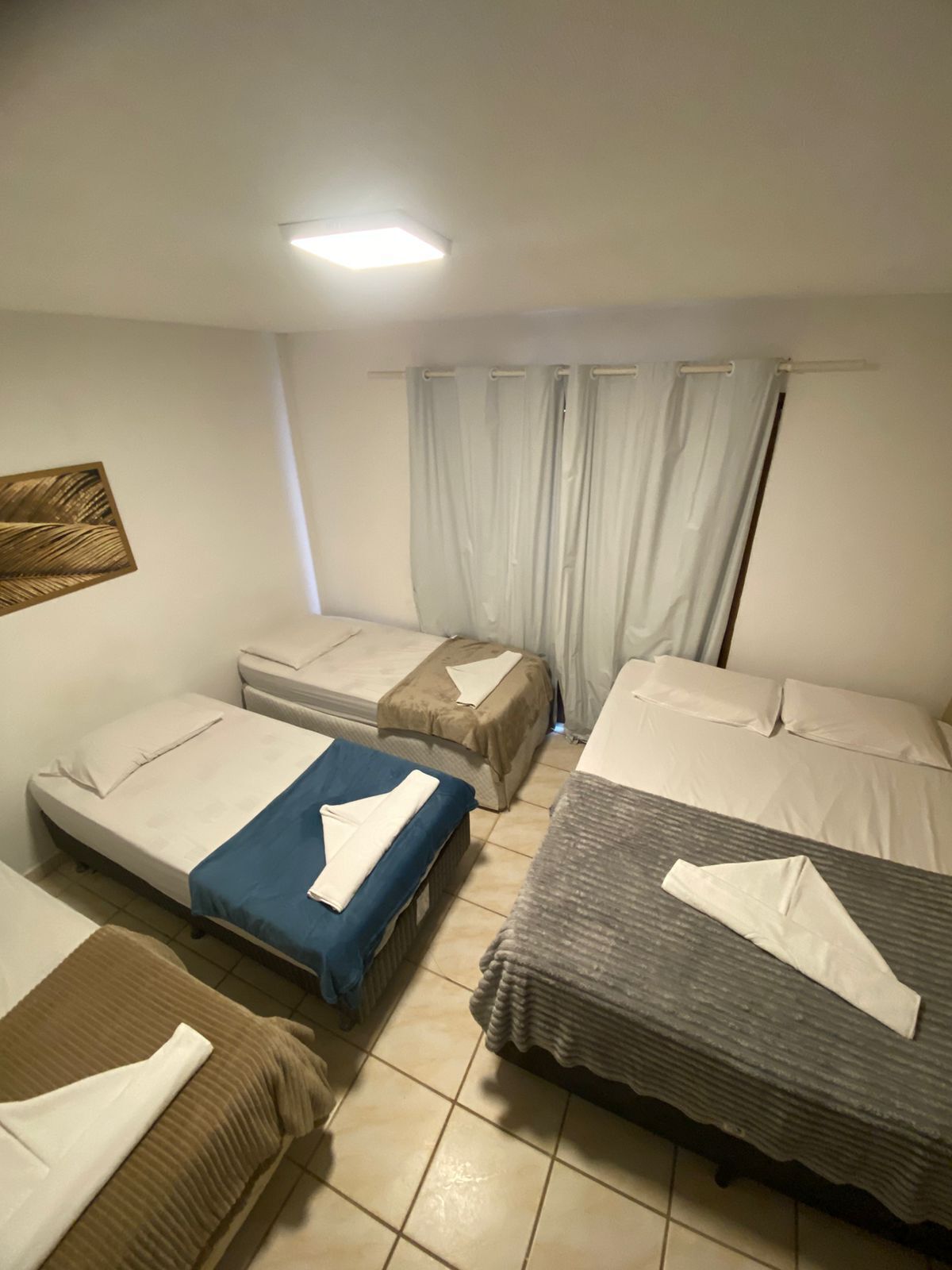 Une chambre avec trois lits, chacun recouvert d’une couvre-lit d’une couleur différente. On y trouve un luminaire au plafond, une fenêtre avec des rideaux et une œuvre d’art accrochée au mur.