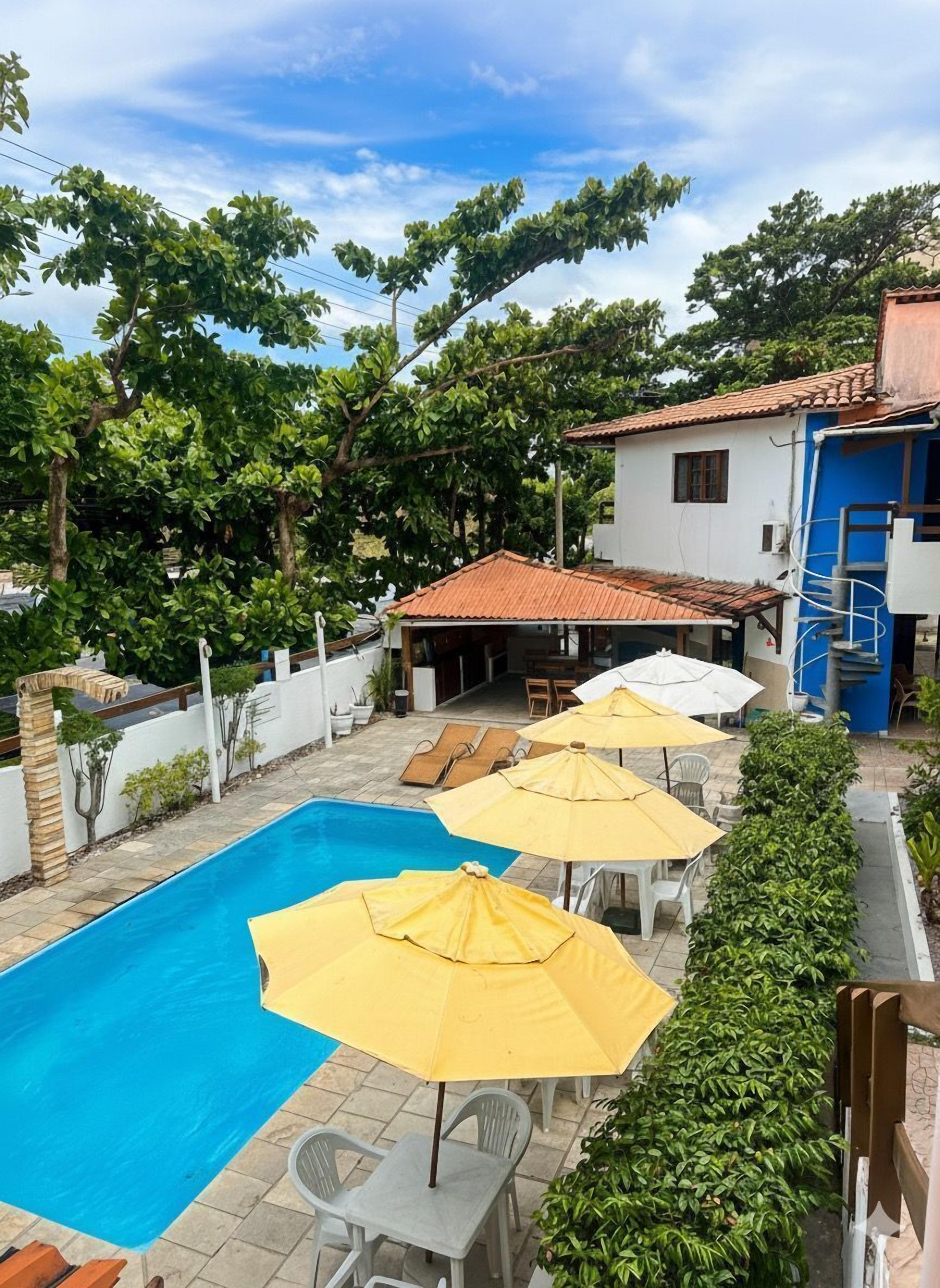 A swimming pool with lounge chairs and umbrellas, surrounded by greenery and a small building with a tiled roof.