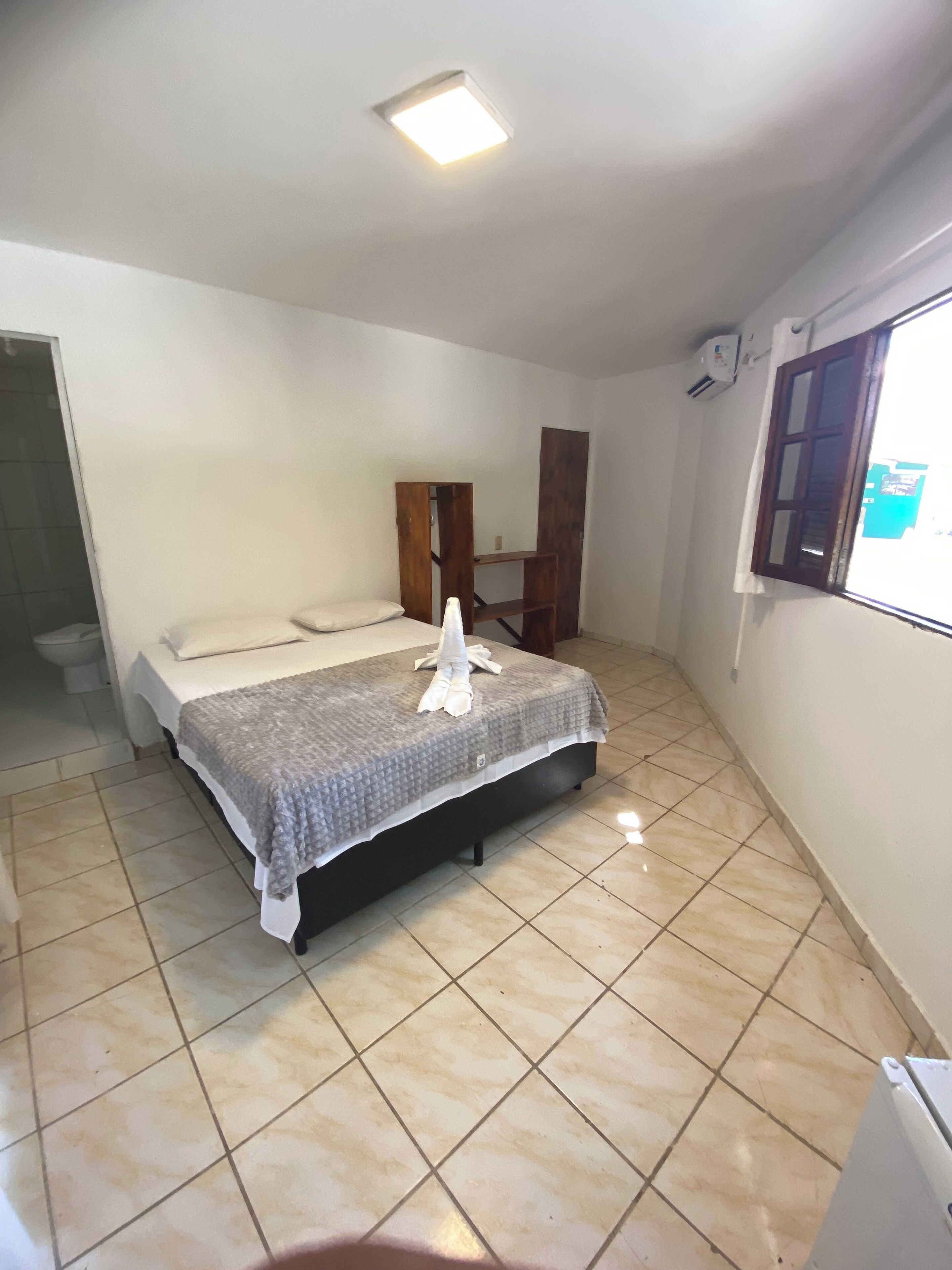 A bedroom with a bed, a wooden wardrobe, a window, and a tiled floor.