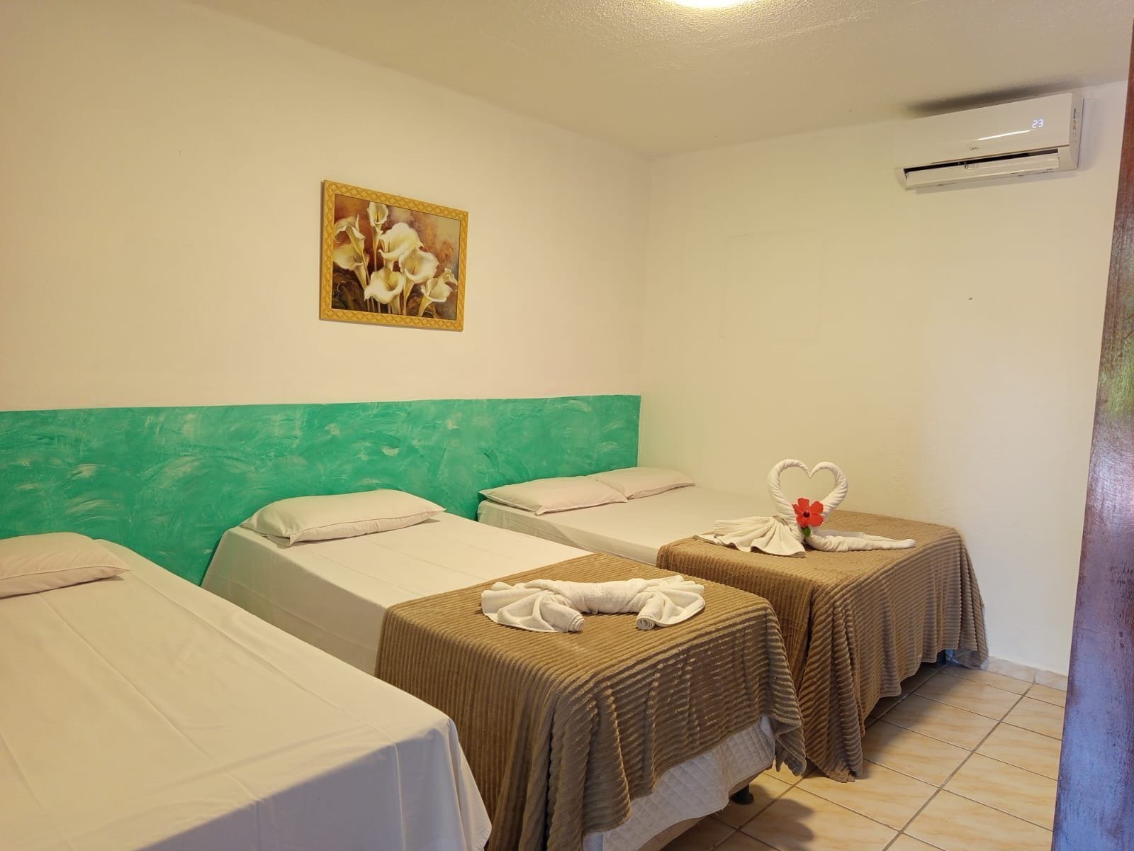 A hotel room with two single beds, a green headboard, a painting of flowers, and an air conditioning unit.
