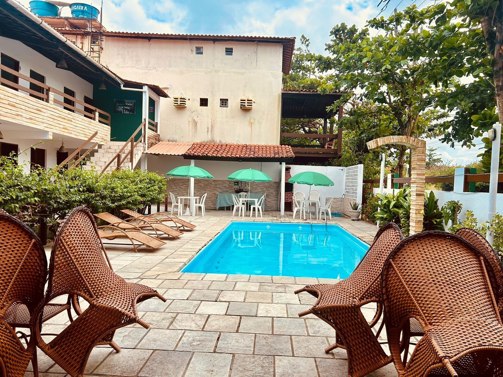 Una piscina con sillones y sombrillas, rodeada por un edificio con techo de tejas y una escalera.