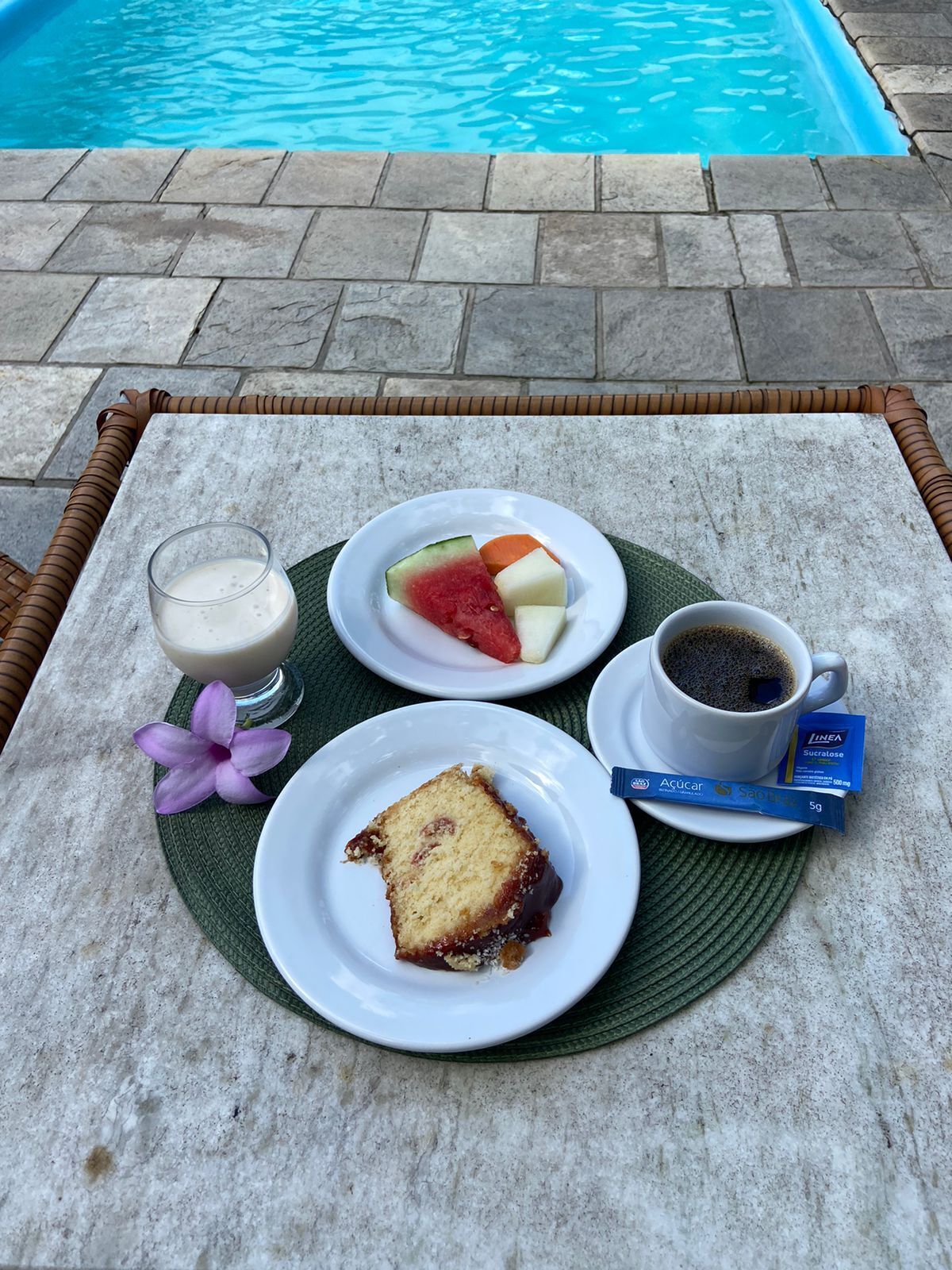 A table with a plate of food, a glass of milk, a cup of coffee, and a packet of medication by a pool.