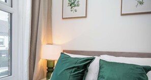 A neatly made bed with green pillows, a bedside lamp, and framed botanical prints on the wall.