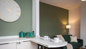 A modern kitchen with a white countertop, a green wall, a clock, and a dining area with a green sofa and a lamp.