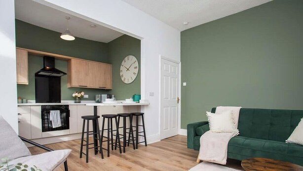A modern living room with a green sofa, a coffee table, and a kitchen area with wooden cabinets and a black range hood. - Vacant Nests | The Jackson | Wi-Fi | Parking (Tynemouth)