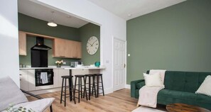 Un salon moderne avec un canapé vert, une table basse et un coin cuisine doté de meubles en bois et d’une hotte noire.