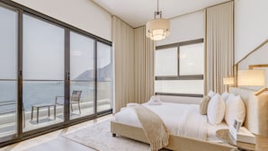 A modern hotel room with a large bed, a view of the sea, and a balcony with a chair.