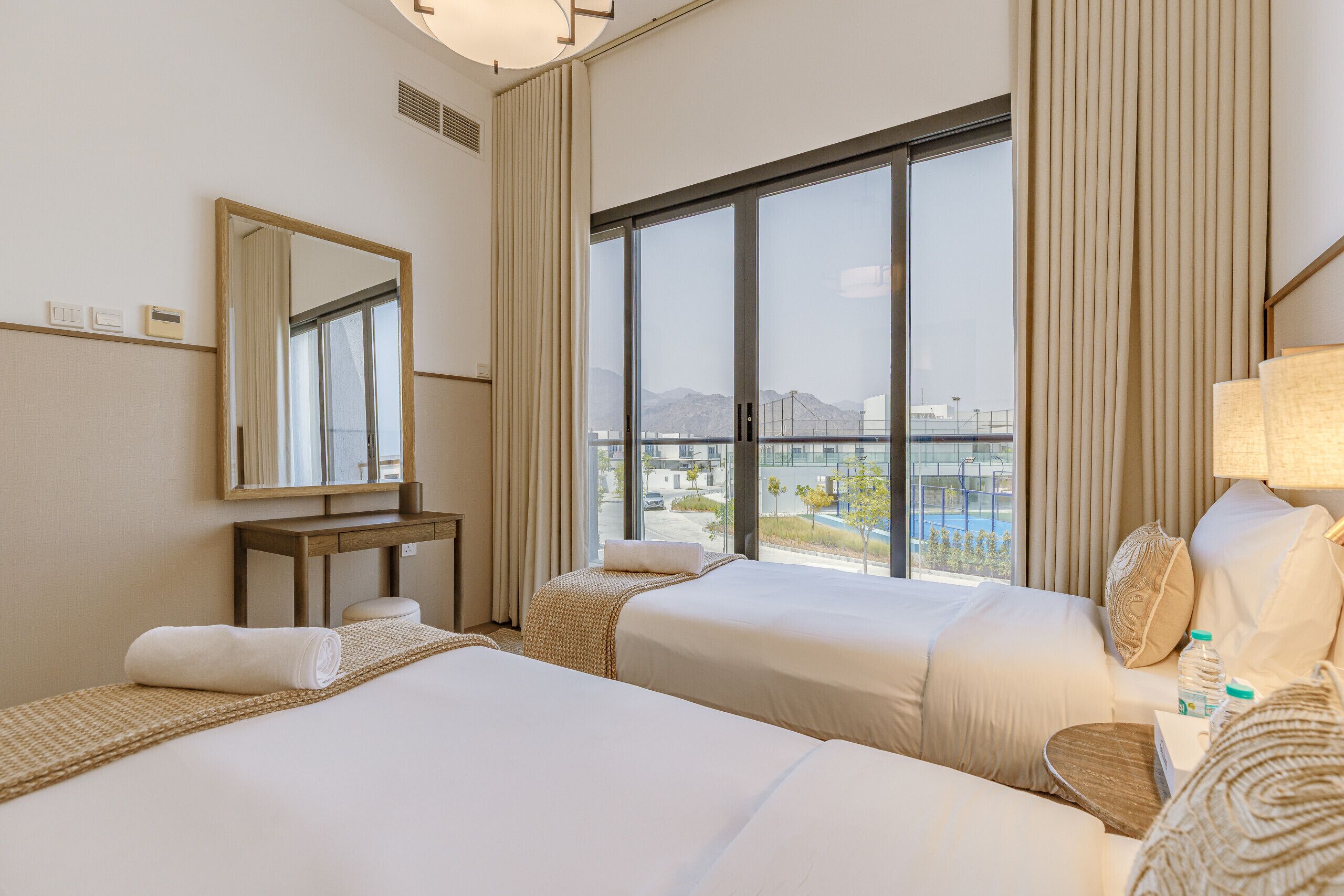 A hotel room with two beds, a large window with a view of a cityscape, a mirror, and a bedside table with a lamp.