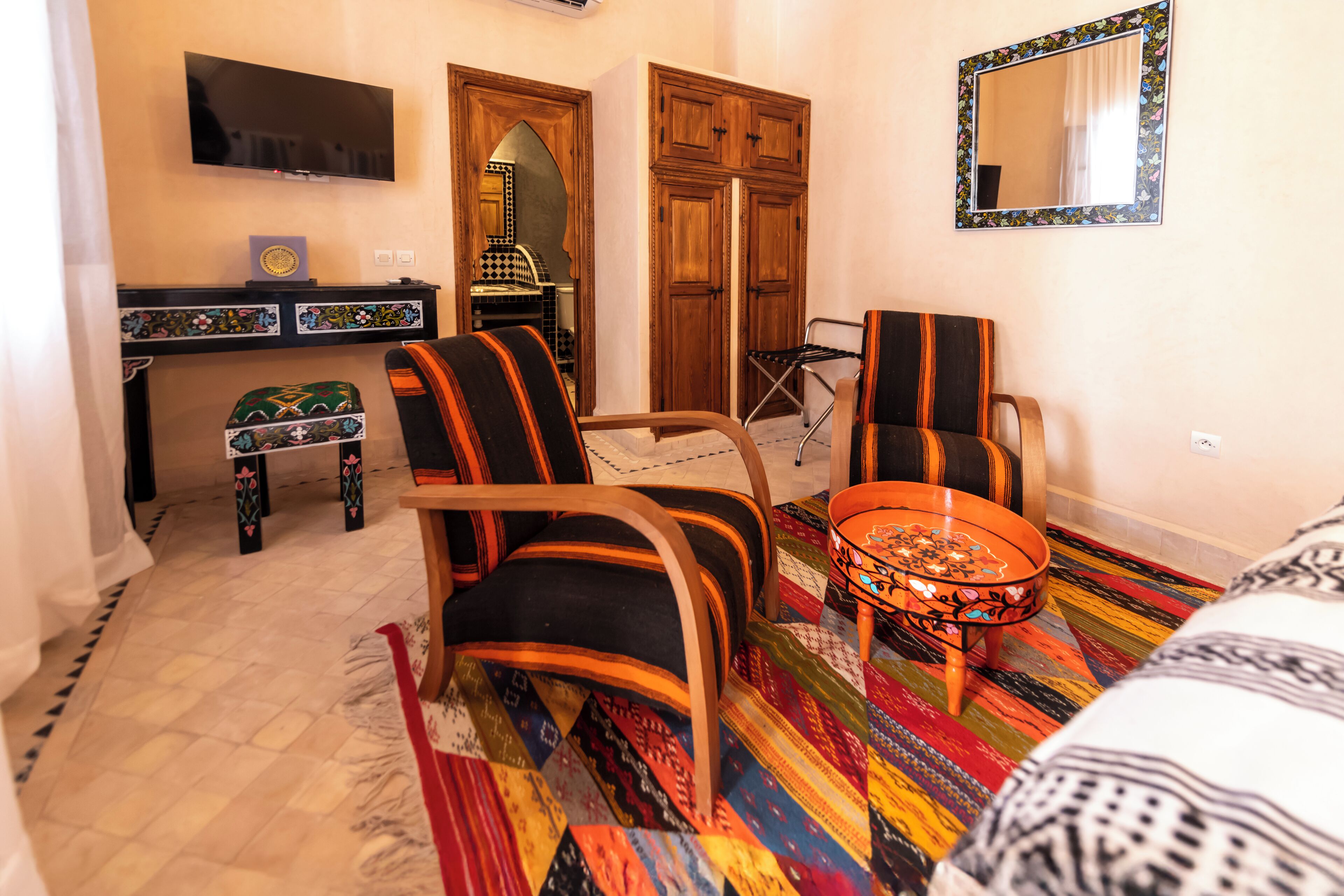 A room with a TV, wooden wardrobe, two chairs, a small table, and a colorful rug.