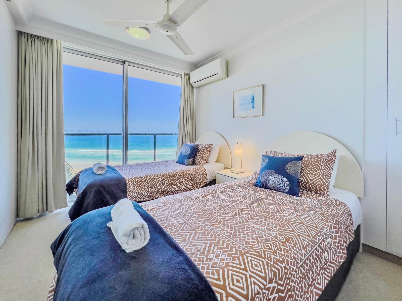 Une chambre d’hôtel avec deux lits, un ventilateur de plafond, une grande fenêtre donnant sur la plage et un tableau encadré au mur.