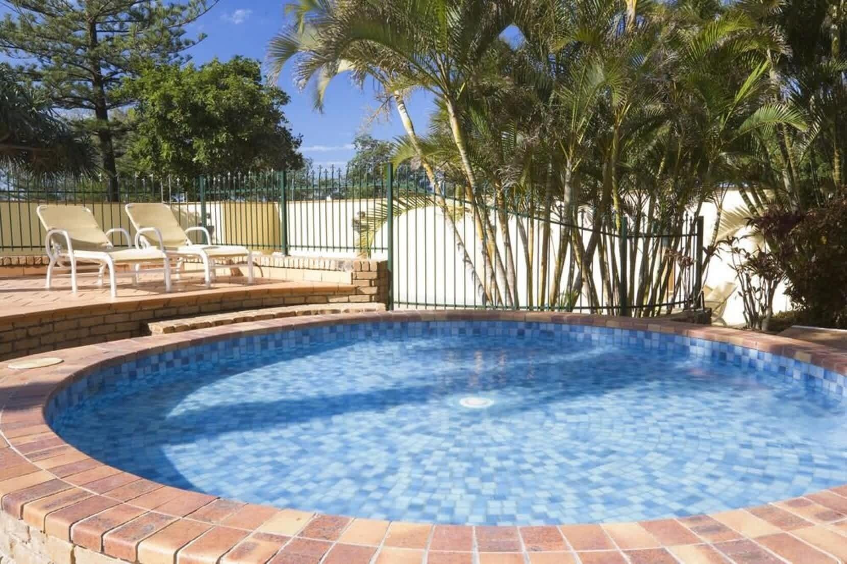 Une piscine dont le bord est revêtu de carrelage, entourée d’une terrasse en briques et d’une clôture en métal.