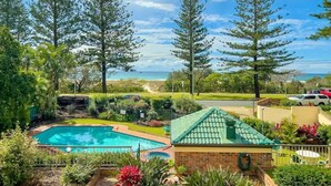 A pool surrounded by lush greenery and tall trees, with a view of the ocean and a clear sky. - Beachfront Apartment with Hot Tub & Pool (Bilinga)