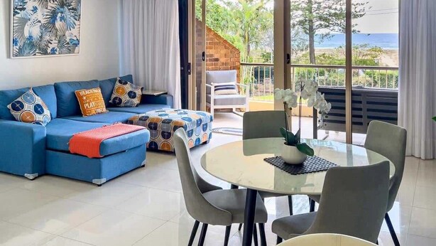 A living room with a blue sofa, a dining table with chairs, and a view of the outdoors through large windows. - Beachfront Apartment with Hot Tub & Pool (Bilinga)