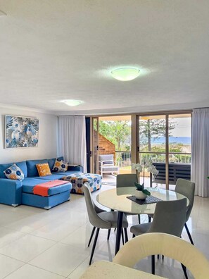 Un salon avec un canapé bleu, une table à manger avec des chaises et une vue sur l’extérieur grâce à de grandes fenêtres.