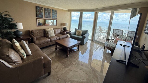 A spacious living room with a large window offering a view of the ocean. There are two sofas, a coffee table, and a TV stand. - Welcome to your perfect beachfront condo in Daytona Beach Shores! (Daytona Beach Shores)