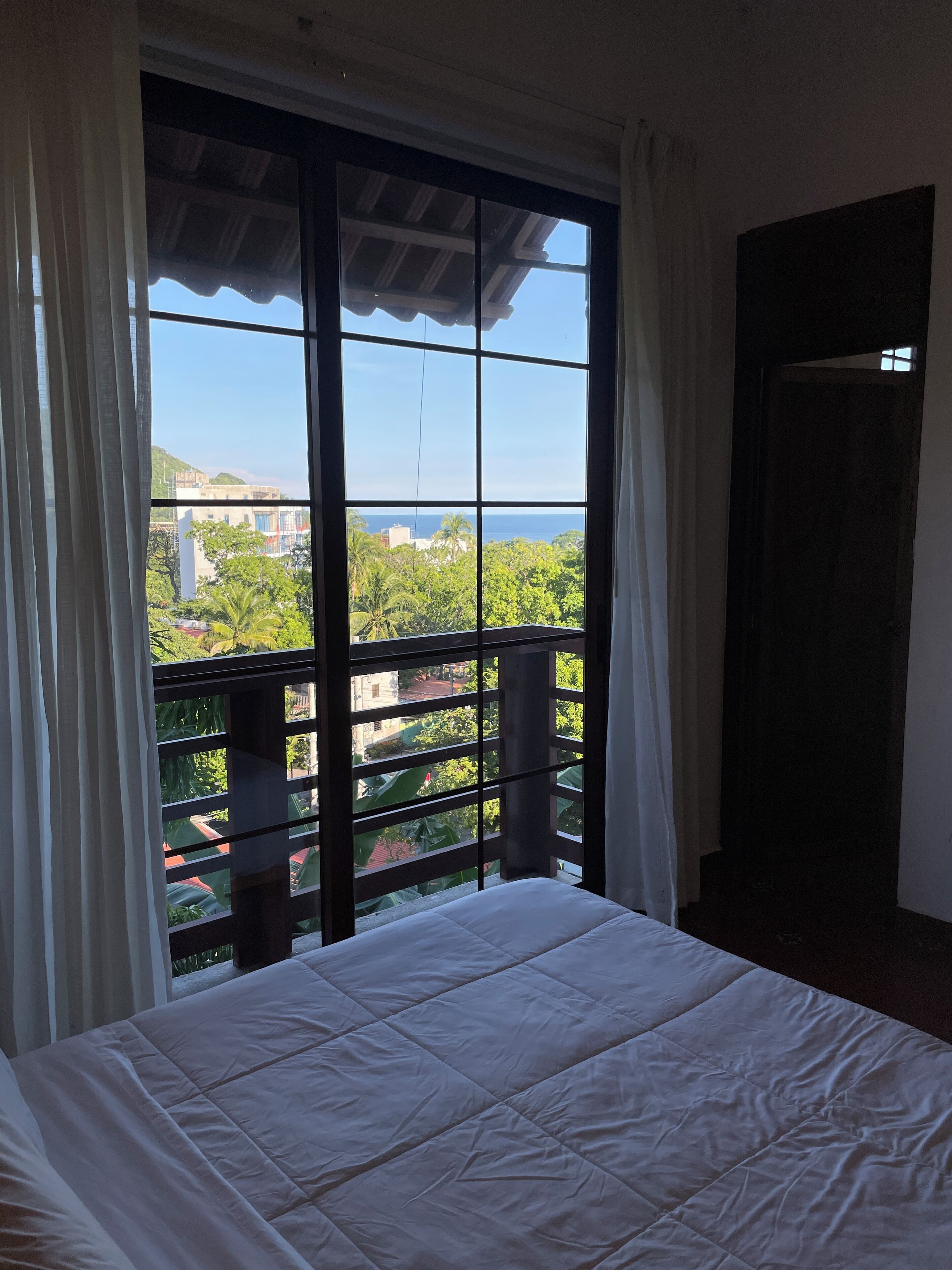 Una habitación con cama, ventanas amplias y vistas a zonas verdes y edificios.