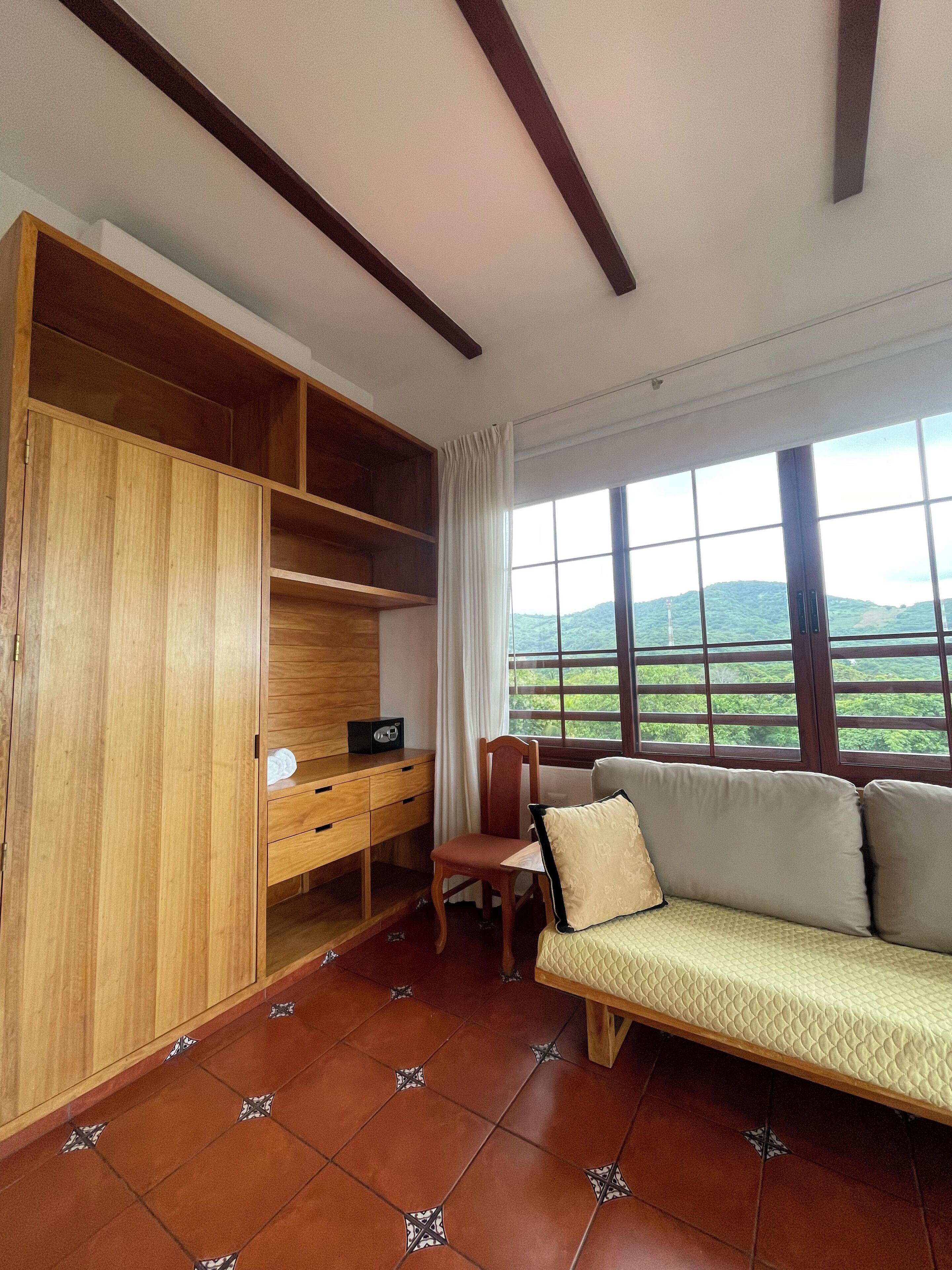 Una habitación con un armario de madera, un escritorio con una radio, una silla, un sofá y un ventanal con vistas a zonas verdes.