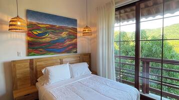 Una camera da letto con una testiera in legno, un letto grande con lenzuola bianche, due lampade a sospensione, un quadro astratto colorato e un'ampia finestra con vista sul verde.