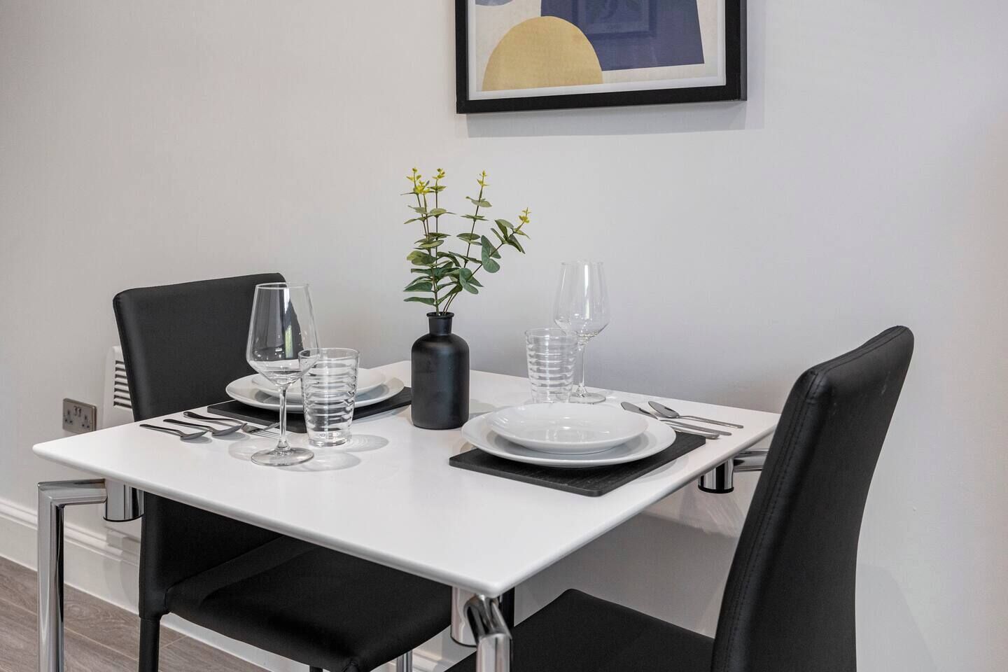 Ein Esstisch ist gedeckt mit Tellern, Besteck und Gläsern und wird von schwarzen Stühlen begleitet. Auf dem Tisch steht eine Vase mit Grünpflanzen.