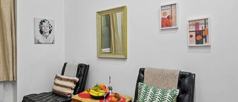 Een woonkamer met een eettafel gedekt met fruit en gebak, twee stoelen, een bank met een groen gekleurd kussen en kunstwerken aan de muur.
