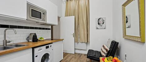 A compact kitchen with a microwave, washing machine, and sink. A dining area with a table set for two, featuring fruits, a croissant, and a glass.