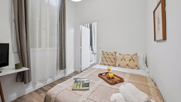 A modern hotel room with a bed, a bedside table, a TV, and a window with sheer curtains.