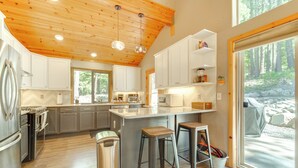 A modern kitchen with wooden ceiling, white cabinets, stainless steel appliances, and a large window overlooking a forest.