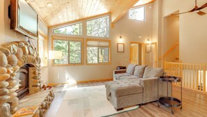 A cozy living room with a stone fireplace, a large window, a grey sofa, and a wooden ceiling.