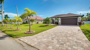 A residential driveway leading to a house with a garage, surrounded by well-maintained lawns and palm trees. - Family Retreat w/ Backyard Oasis in Cape Coral! (Cape Coral)