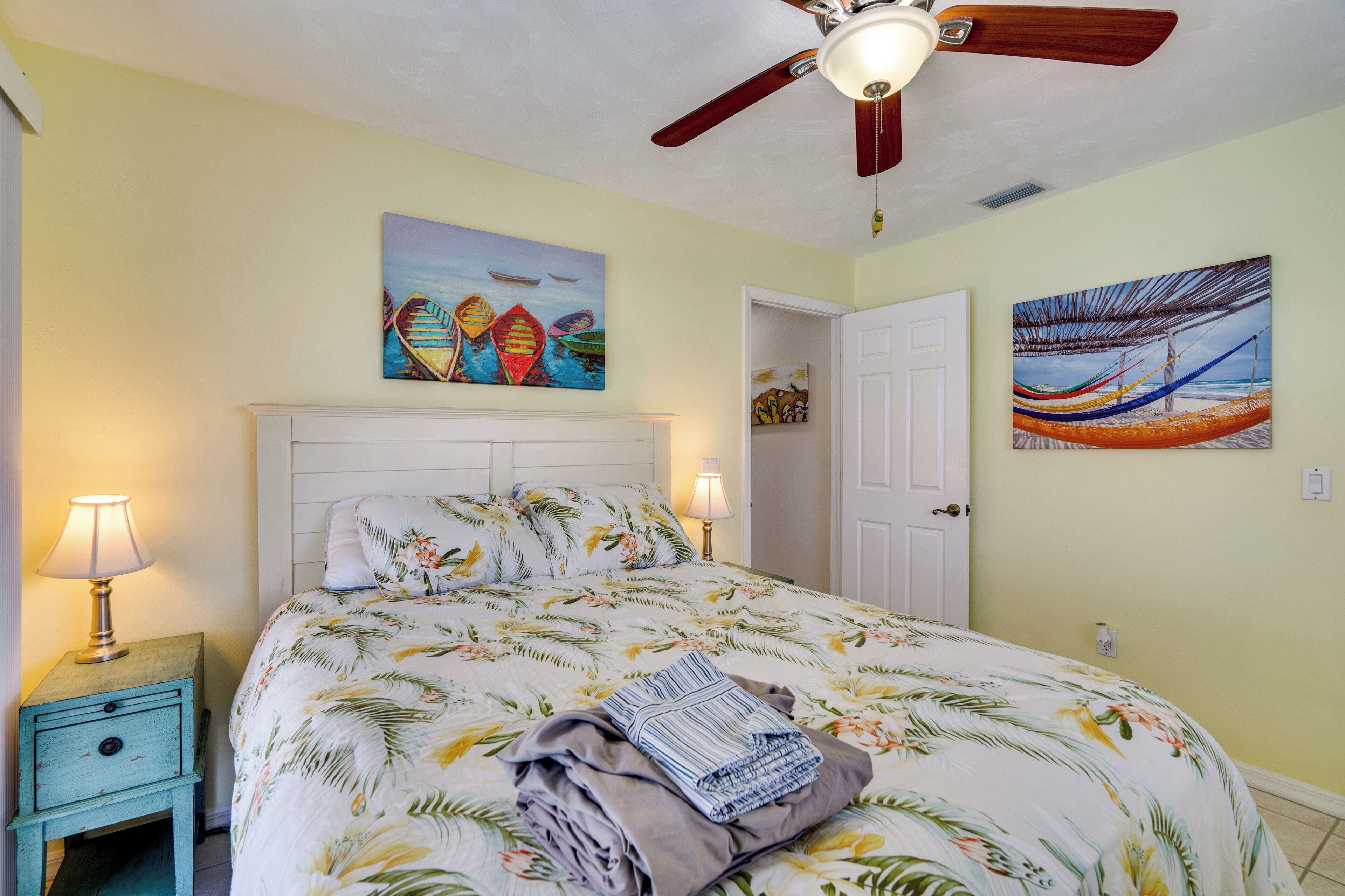 A bedroom with a bed, a nightstand, a ceiling fan, and two paintings on the walls.