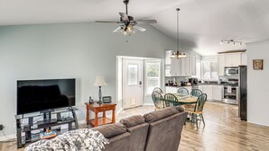 A modern living room with a sofa, TV, and ceiling fan. A dining area with chairs and a table is visible. The kitchen has white cabinets and appliances. - Family Home w/ Fenced Yard Between Provo & Slc! (Herriman)