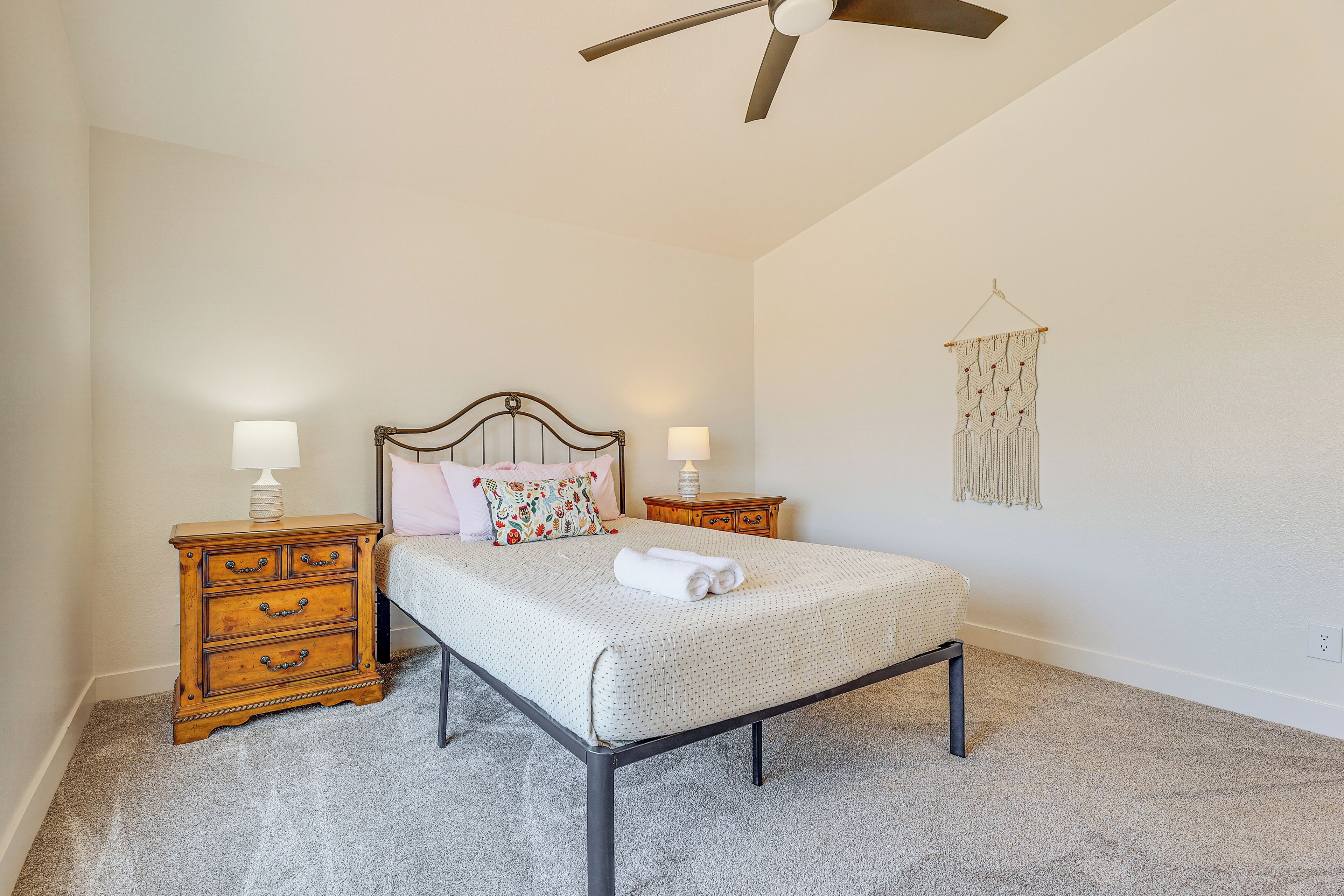A bedroom with a bed, two bedside tables, a ceiling fan, a wall hanging, and a carpeted floor.