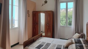 Un dormitorio con cama, un armario de madera, un ventilador de techo y dos ventanas con cortinas.
