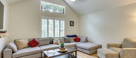 A spacious living room with a high ceiling, a large window with blinds, a ceiling fan, and a sectional sofa with decorative pillows.