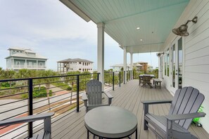 Eine überdachte Terrasse im Freien mit Adirondack-Stühlen und einem Tisch, mit Blick auf einen Strand mit Häusern im Hintergrund.