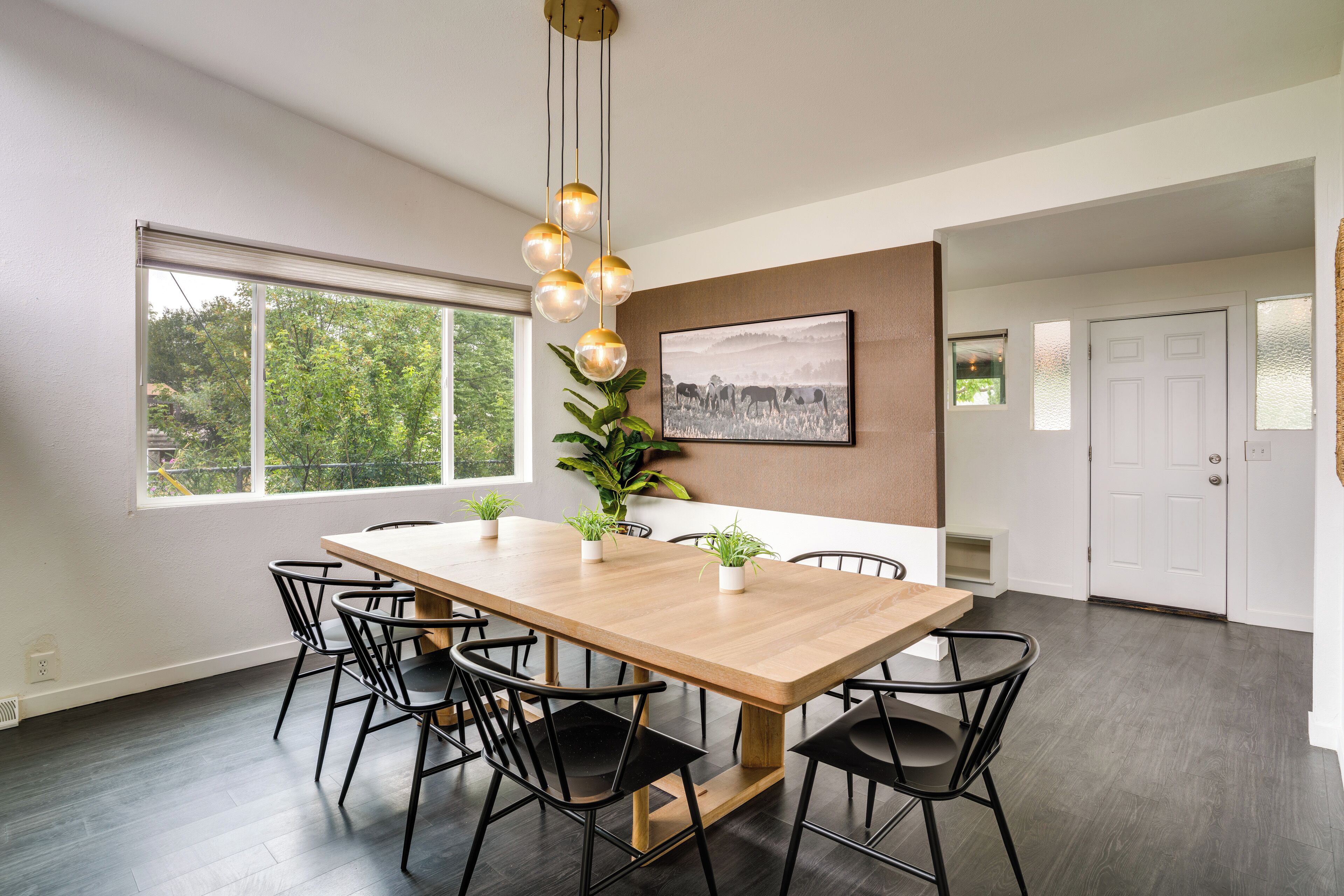 A modern dining area with a wooden table and black chairs, large windows, and a framed picture on the wall.