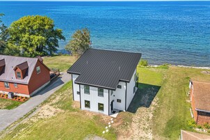 Uma casa moderna de dois andares com telhado plano, localizada em uma área gramada com vista para uma grande massa de água.