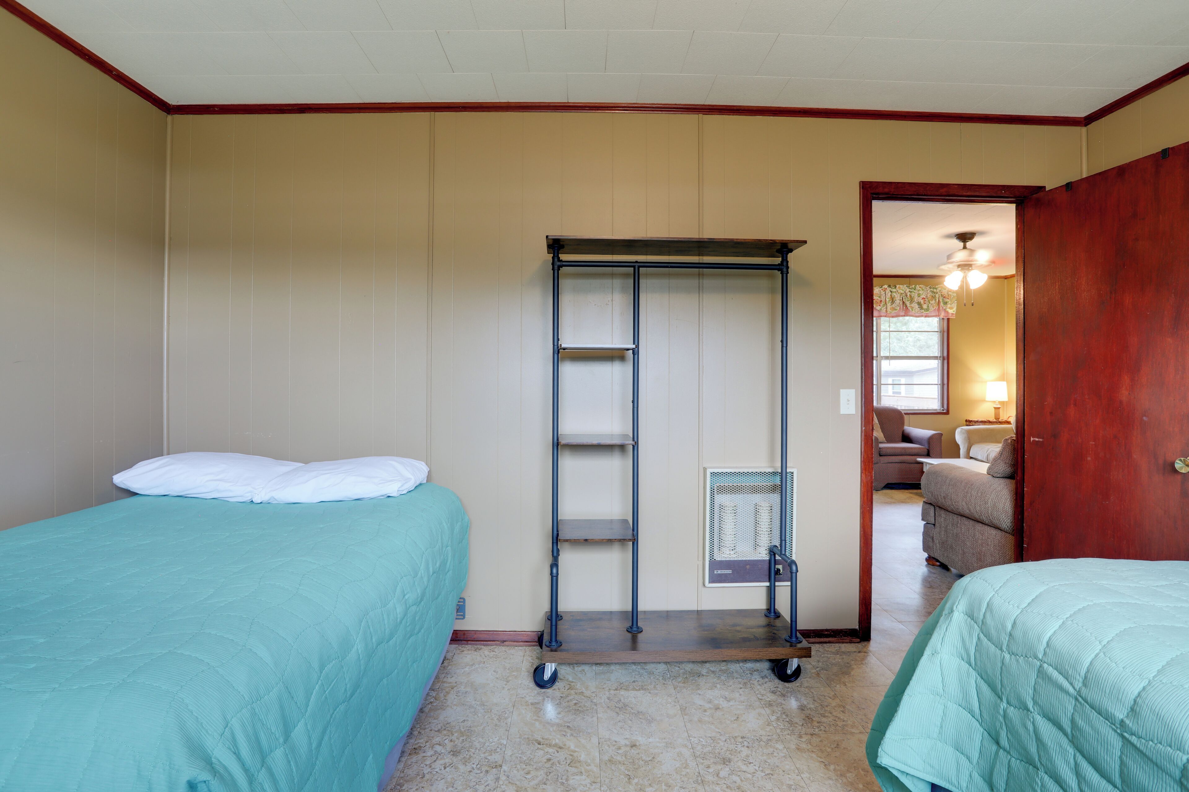 A room with two beds, a metal shelf unit, and a doorway leading to another room with a chair and lamp.