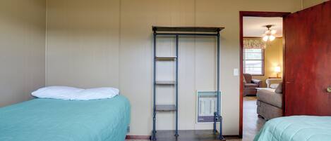A room with two beds, a metal shelf unit, and a doorway leading to another room with a chair and lamp.