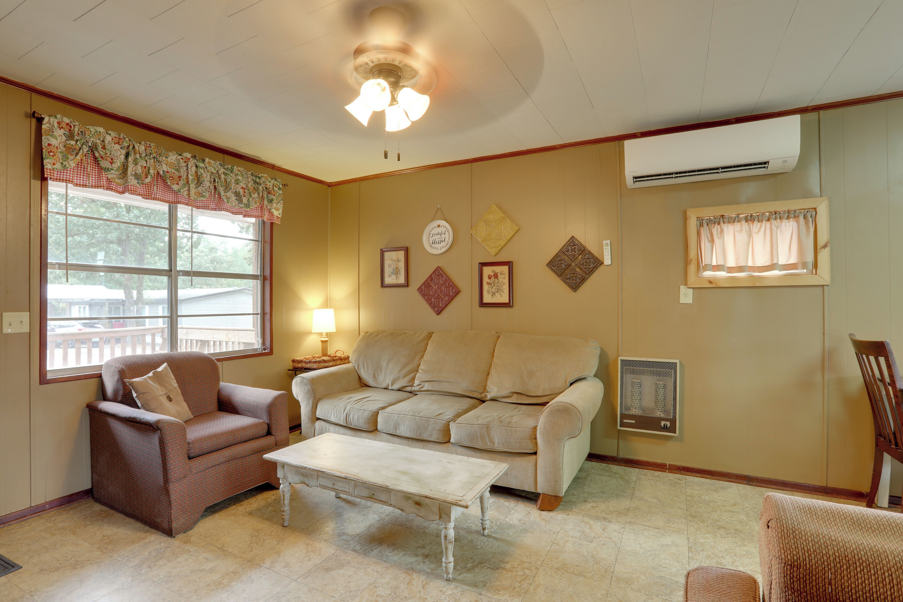 A living room with a beige sofa, a brown armchair, and a white coffee table. There is a window with a floral valance, a ceiling fan, and wall-mounted decorations.
