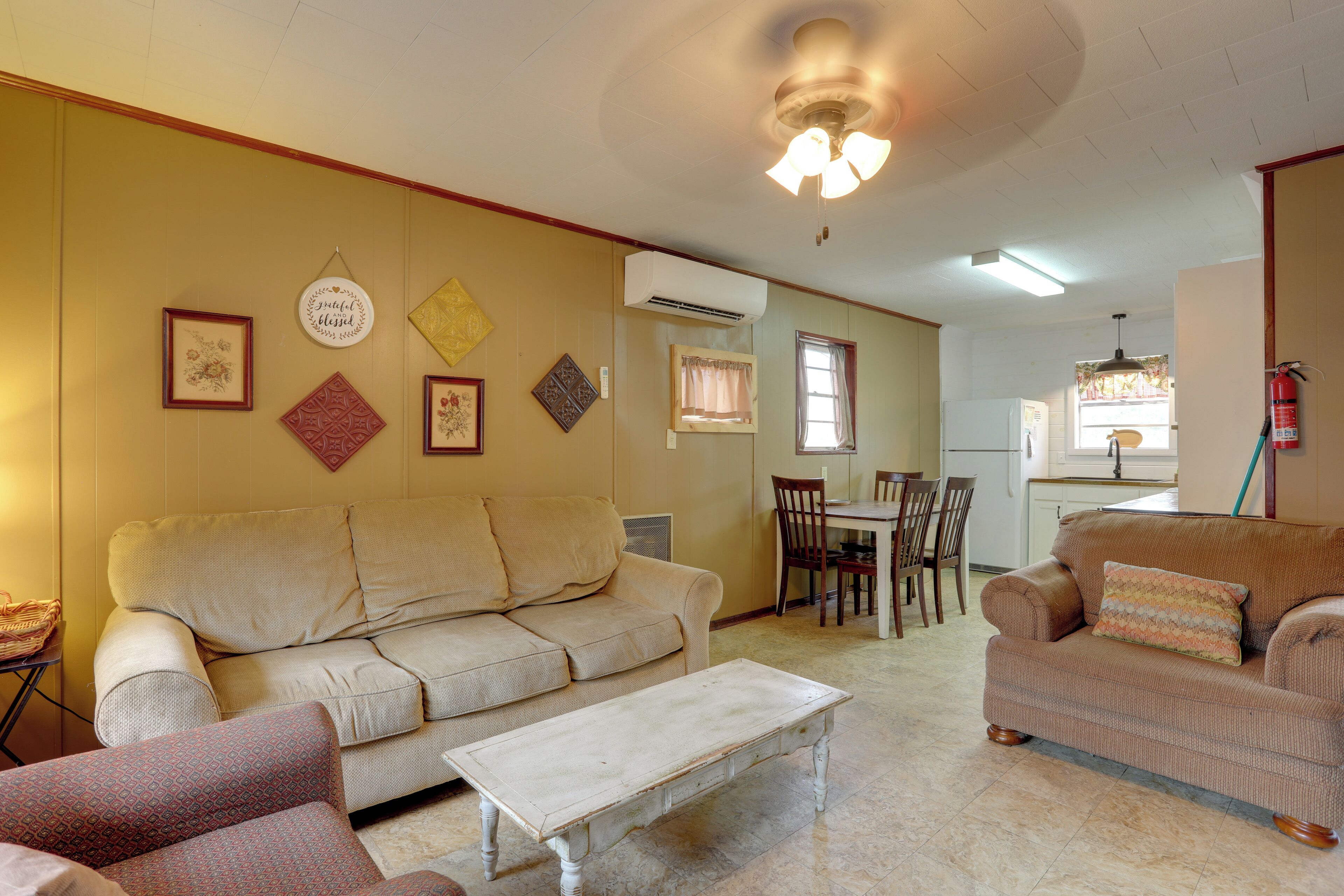 Una sala de estar con un sofá color beige, un sillón estampado y una mesa de centro blanca. Se observa una zona de comedor con una mesa y sillas al fondo.