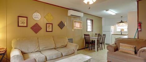 Una sala de estar con un sofá color beige, un sillón estampado y una mesa de centro blanca. Se observa una zona de comedor con una mesa y sillas al fondo.