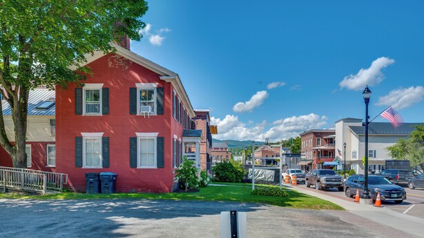 A red building with white shutters, a flagpole with an American flag, and a street with parked cars and traffic cones. - Waterbury Studio 12 Mi to Bolton Valley Ski Resort (Waterbury)