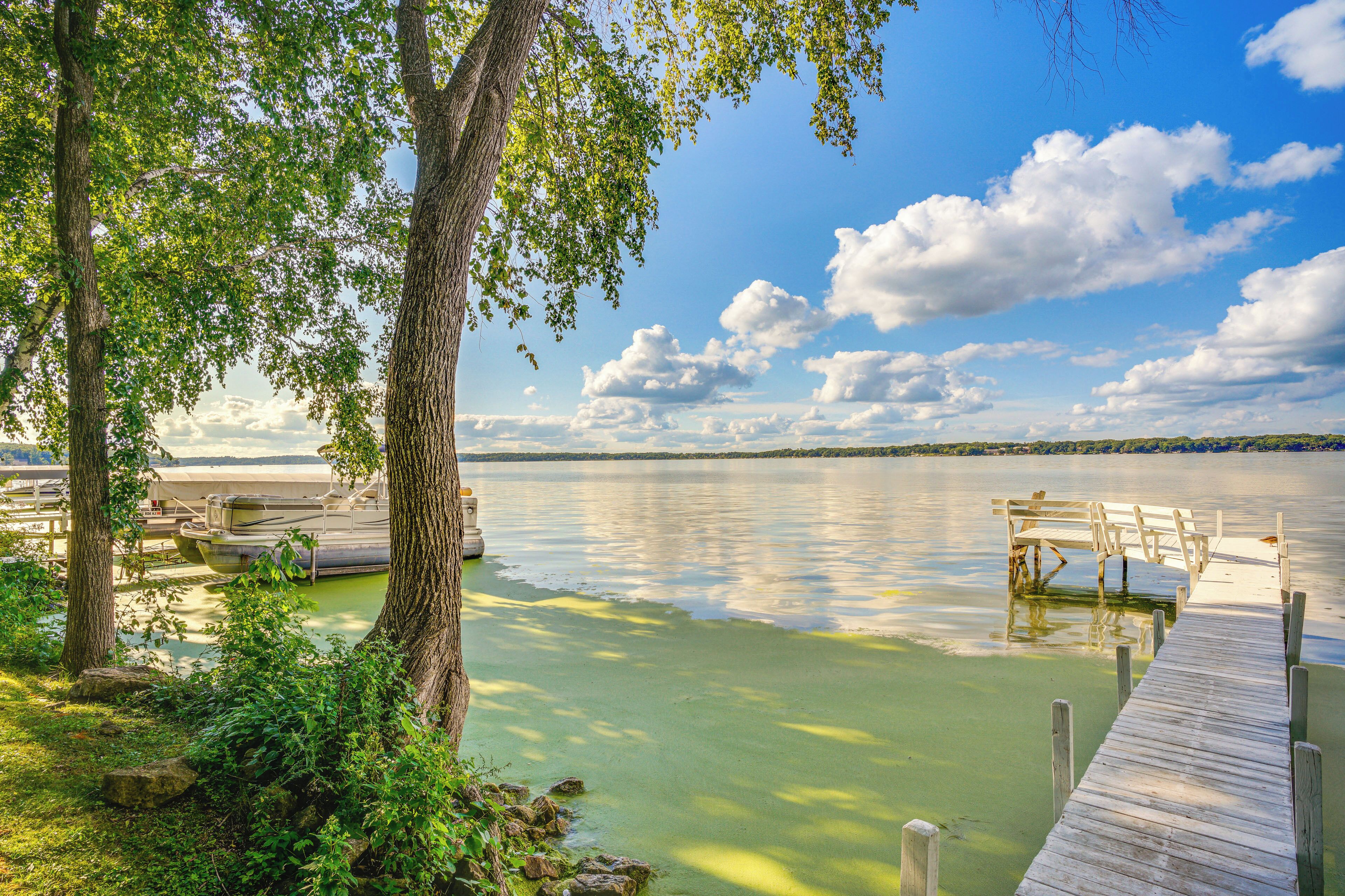 A wooden dock extending into a calm lake with boats docked, surrounded by trees and greenery.