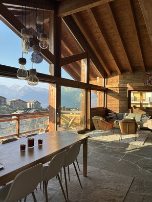 Una habitación amplia con techo de madera, ventanas grandes que ofrecen vistas a la montaña, una zona de comedor con mesa y sillas, y un rincón de estar acogedor con sofá y sillones.