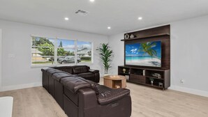 Een moderne woonkamer met een hoekbank, een flatscreen televisie bevestigd aan een houten wand en een groot raam met zonwering.