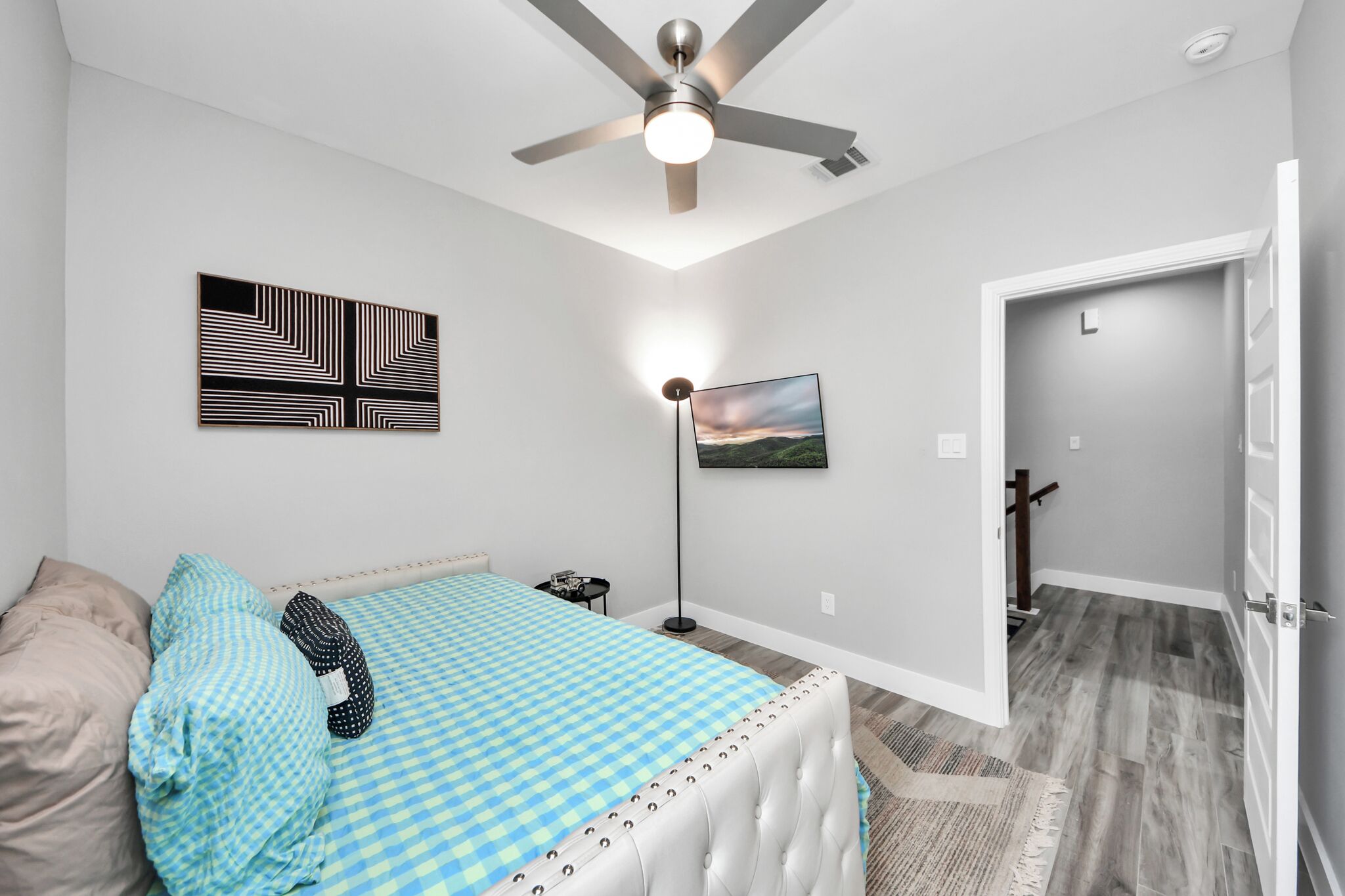 A bedroom with a bed, a ceiling fan, a floor lamp, a painting, and a door leading to another room.