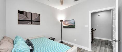 A bedroom with a bed, a ceiling fan, a floor lamp, a painting, and a door leading to another room.