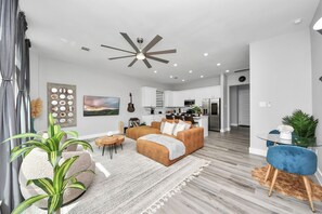 A modern living room with a sofa, coffee table, and a dining area with a glass table and blue chairs. - Private Pool at Pershing Pad. 3BR Sleeps 10. 10 mins from NRG . Huge backyard (Houston)